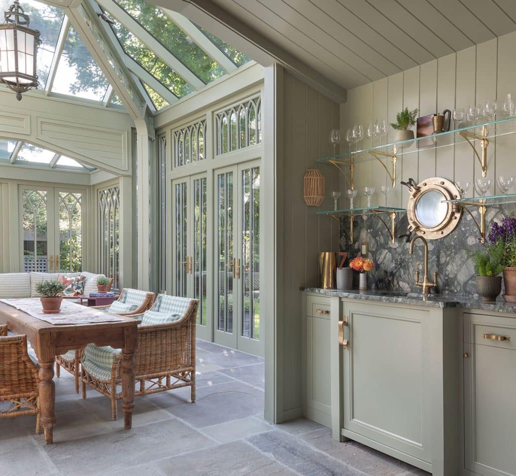 A bright, elegant glass conservatory kitchen with large windows, a glass ceiling, wicker chairs around a wooden table, and a sage green kitchenette with open shelves, glasses, and plants, overlooking the garden.