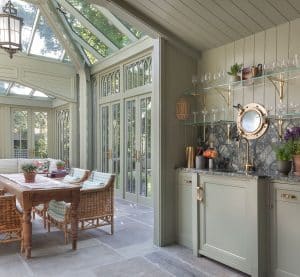A bright, elegant glass conservatory kitchen with large windows, a glass ceiling, wicker chairs around a wooden table, and a sage green kitchenette with open shelves, glasses, and plants, overlooking the garden.