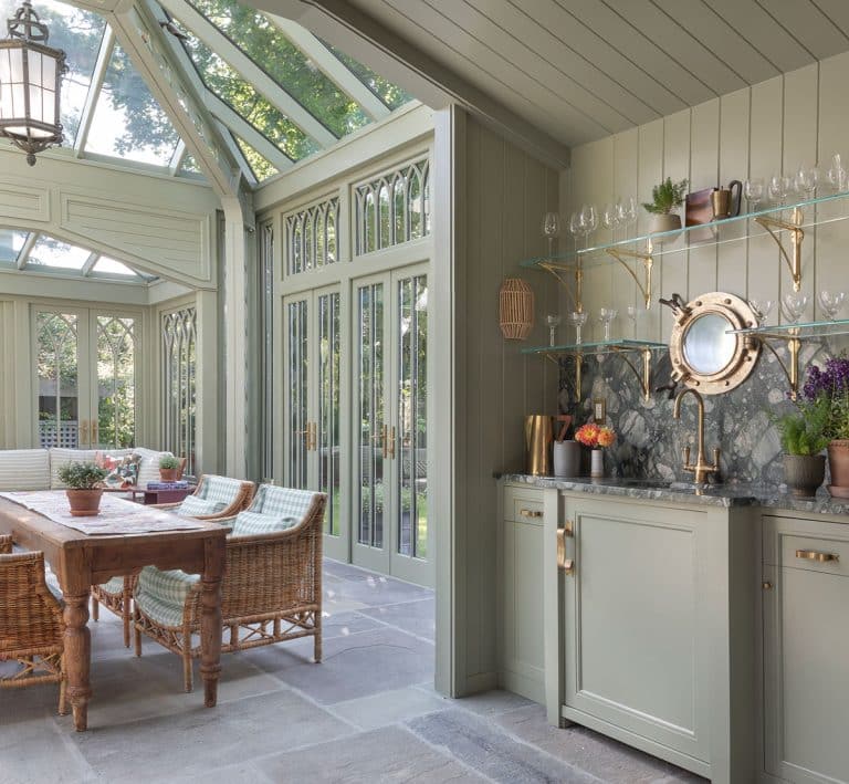 A bright, elegant glass conservatory kitchen with large windows, a glass ceiling, wicker chairs around a wooden table, and a sage green kitchenette with open shelves, glasses, and plants, overlooking the garden.