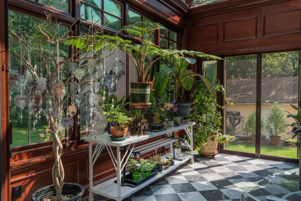 A sunlit glass room with large windows, a checkered black-and-white floor, and various potted plants on white metal shelves. Sunlight streams in, highlighting the lush greenery and offering views typical of elegant glass conservatories.