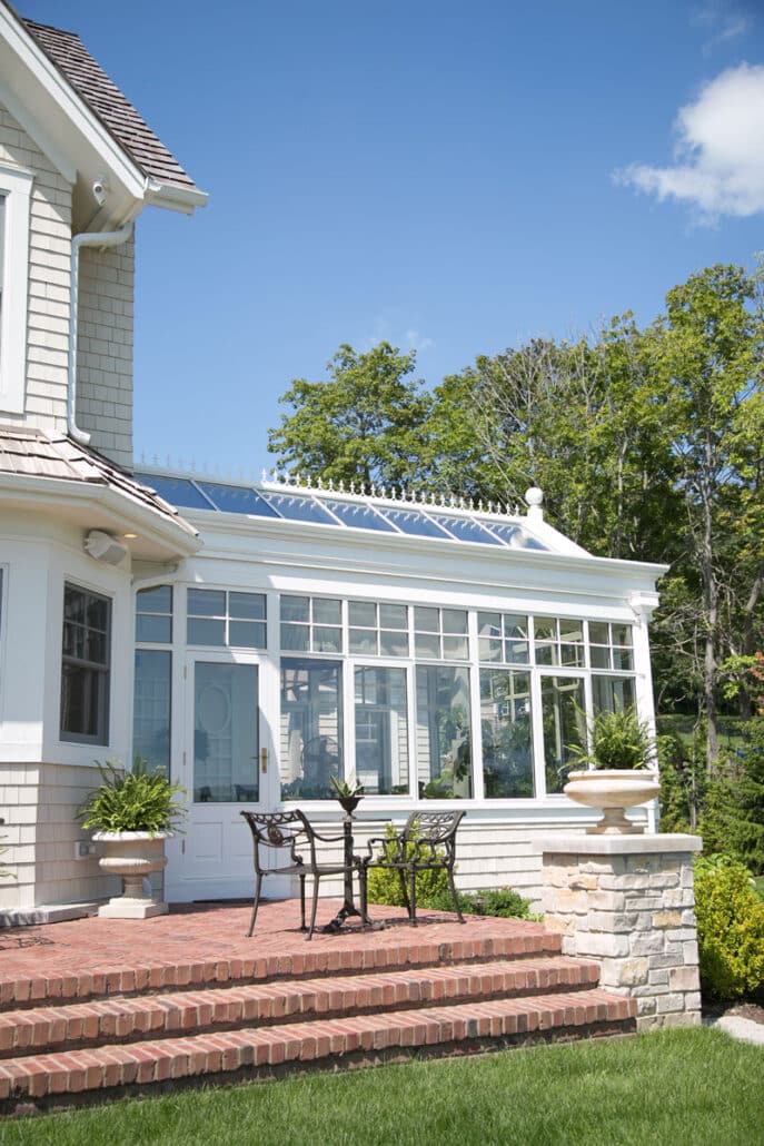 A bright sunroom with large windows and a glass roof extends from a white house, showcasing elegant residential structures. In front, a small metal table with two chairs sits on a brick patio surrounded by greenery.