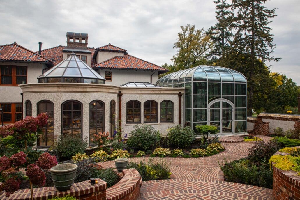A large residential structure with white stucco walls and a red tile roof features a glass conservatory and arched windows. Brick pathways wind through lush, landscaped gardens in the foreground.