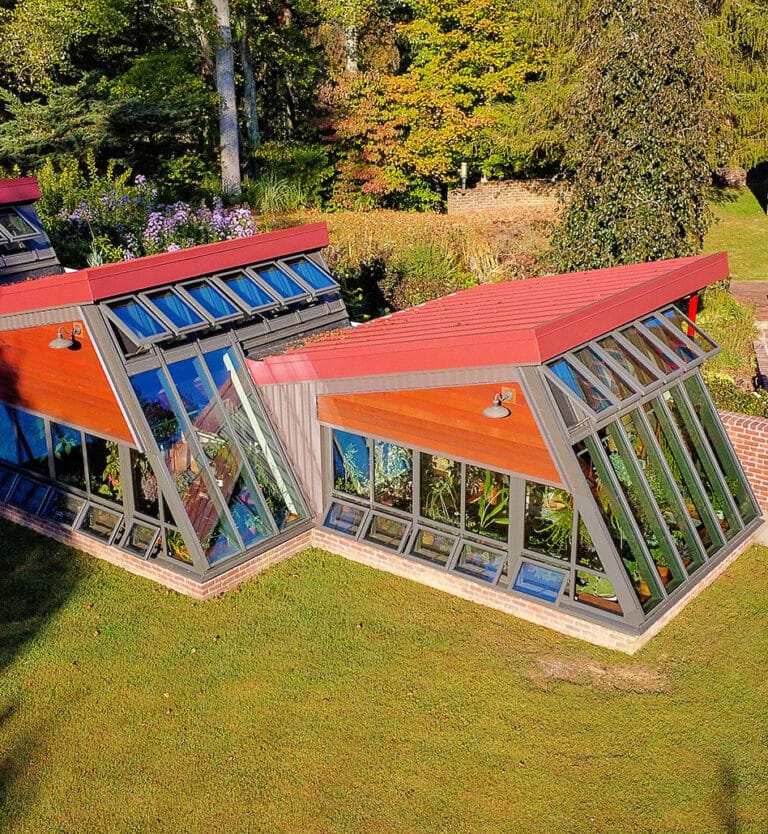 A modern greenhouse with angled glass walls and red roofs stands on a grassy lawn, blending seamlessly with other residential structures, surrounded by trees and garden plants under bright sunlight.