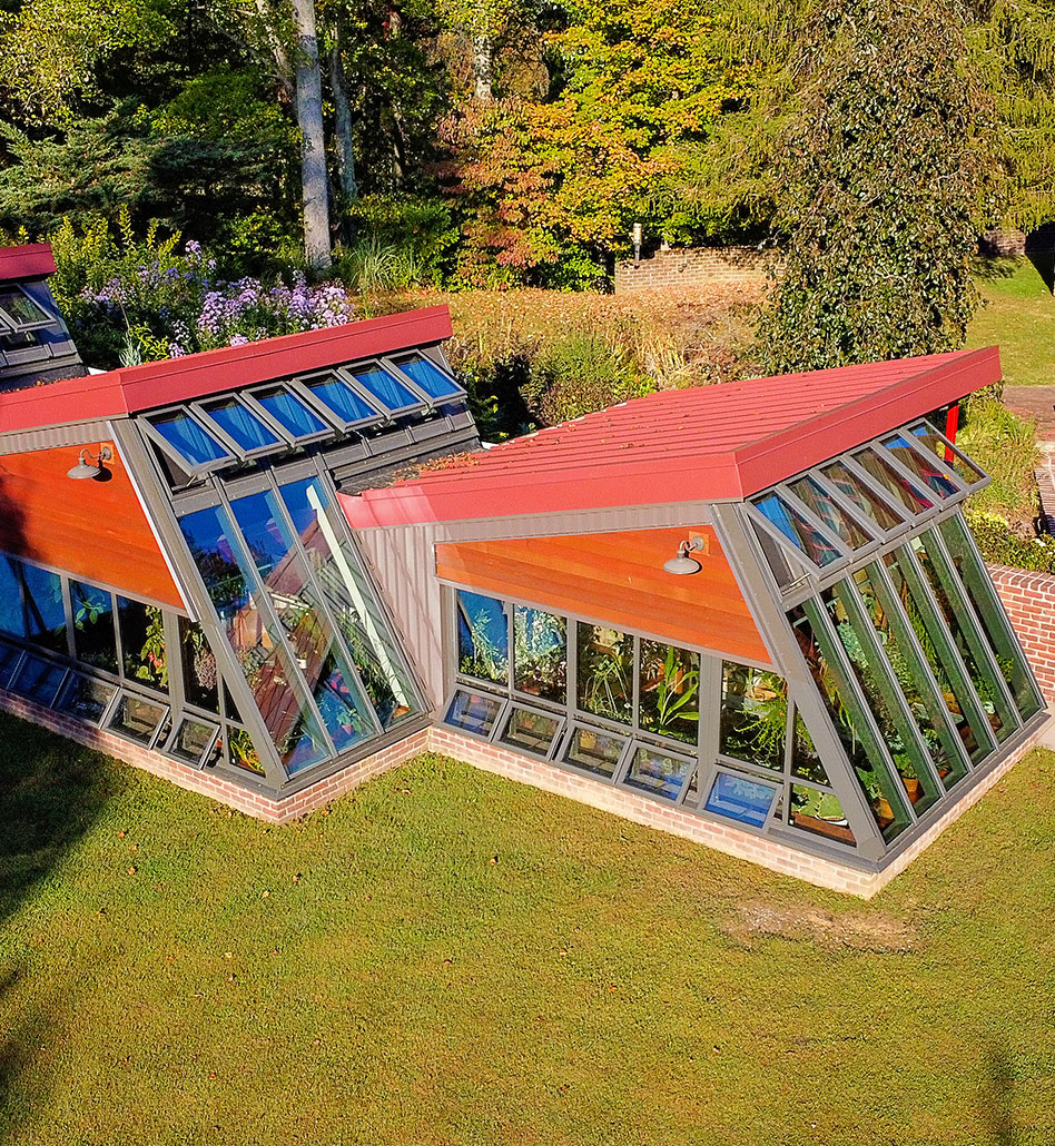 A modern greenhouse with angled glass walls and red roofs stands on a grassy lawn, blending seamlessly with other residential structures, surrounded by trees and garden plants under bright sunlight.