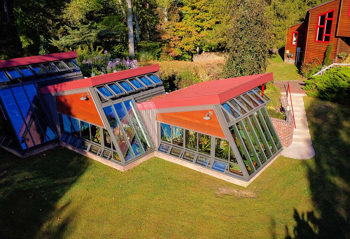 Aerial view of a modern greenhouse with slanted glass walls and red roofs, surrounded by grass and trees, next to a sidewalk leading to a red house.