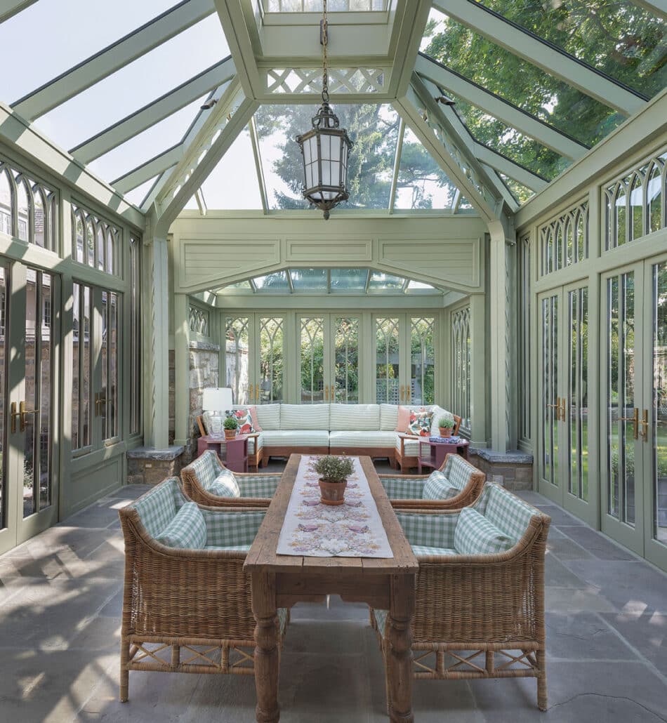 A bright, glass-roofed sunroom—one of the most inviting residential structures—with wicker chairs, a wooden table, and cushioned benches. Large windows and doors surround the space, letting in sunlight and views of greenery outside.