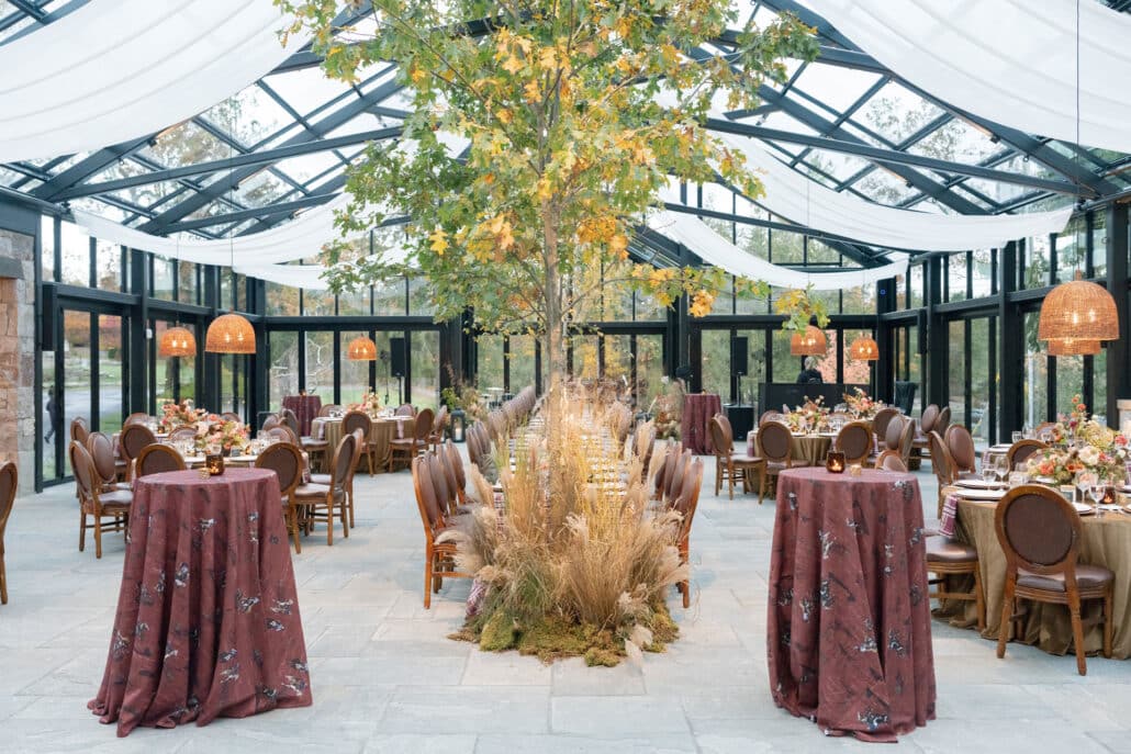 Elegant indoor event with a long dining table centered under a tall tree, surrounded by round tables and high cocktail tables with maroon cloths, in a glass-ceilinged room with natural light and hanging wicker lamps.