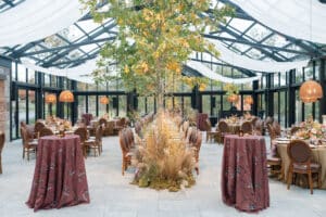 Elegant indoor event with a long dining table centered under a tall tree, surrounded by round tables and high cocktail tables with maroon cloths, in a glass-ceilinged room with natural light and hanging wicker lamps.