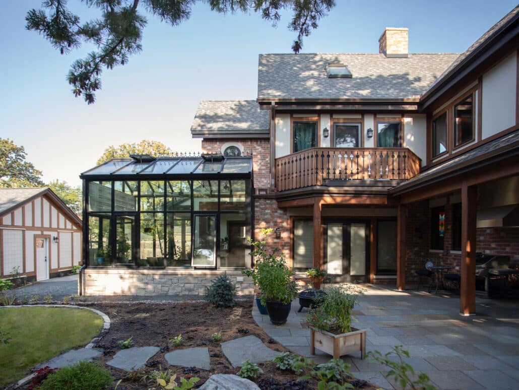 A two-story house with a glass conservatory, brick and wood exterior, balcony, and a landscaped patio area with potted plants and garden beds.