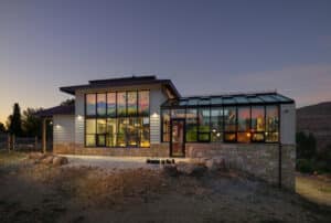 Modern house with large windows and a glass sunroom, set on rocky terrain at dusk. Warm interior lights glow, and a mural of mountains is visible through the main windows. Sky is clear with hints of sunset.