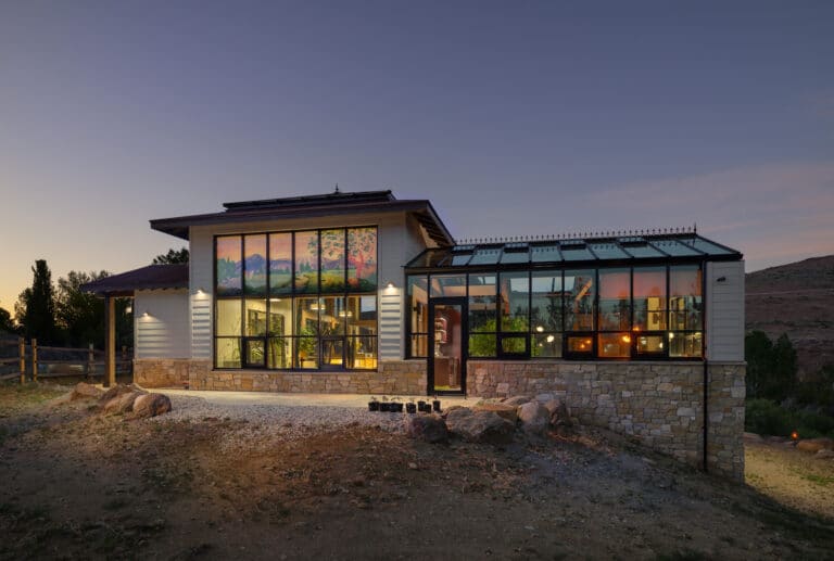 Modern house with large windows and a glass sunroom, set on rocky terrain at dusk. Warm interior lights glow, and a mural of mountains is visible through the main windows. Sky is clear with hints of sunset.