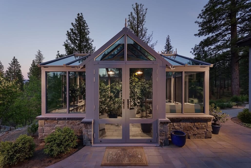 A glass-walled sunroom with a gabled roof set on a stone foundation at twilight, with potted plants visible through the double doors.